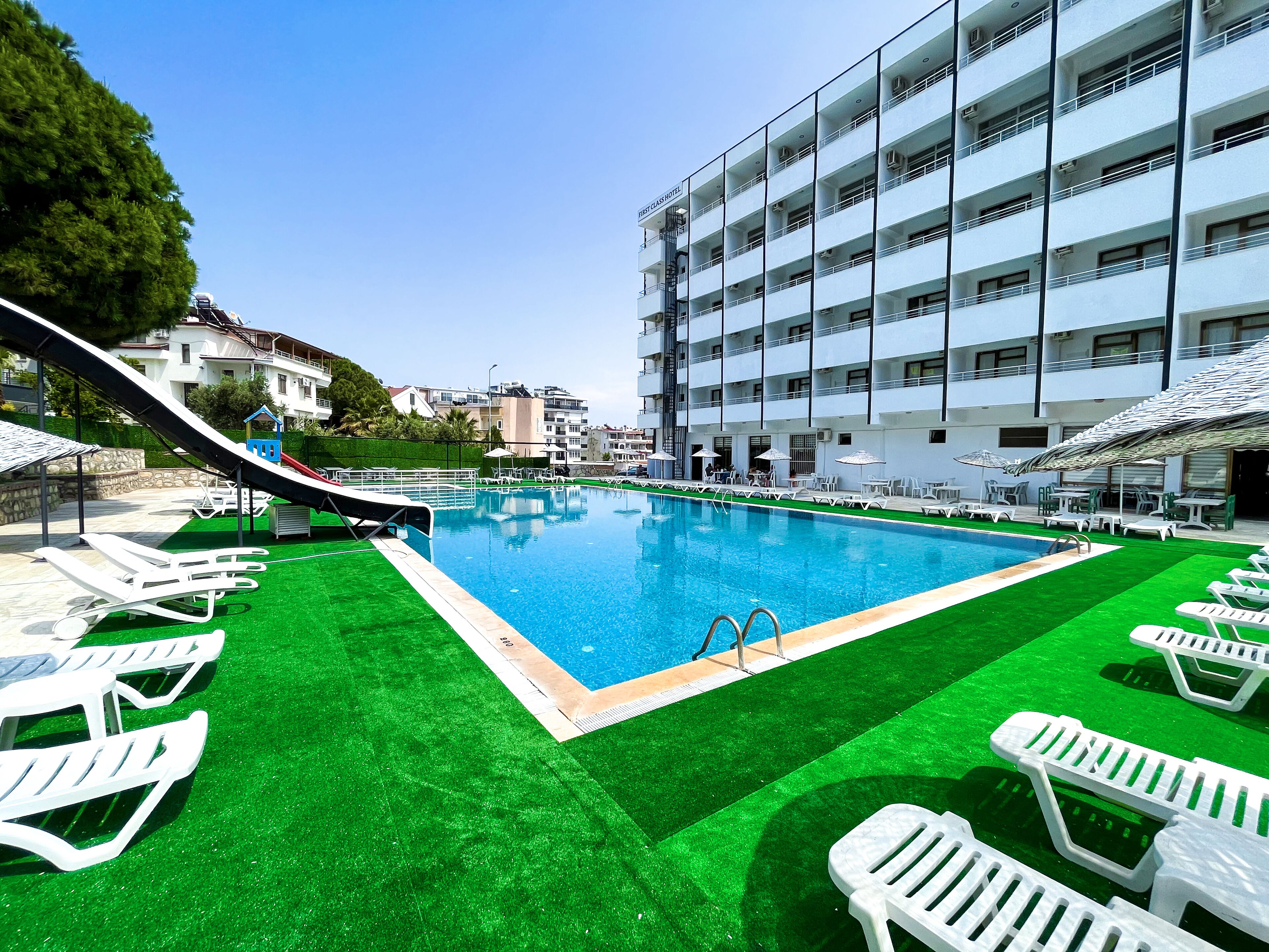 A swimming pool with a slide, surrounded by white lounge chairs and umbrellas, and a building labeled "FIRST CASH HOTEL" in the background.