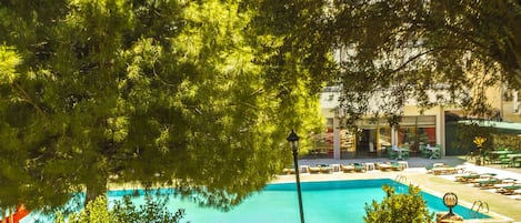 Una piscina con sillas de descanso y sombrillas, rodeada de árboles y un edificio con fachada de vidrio.