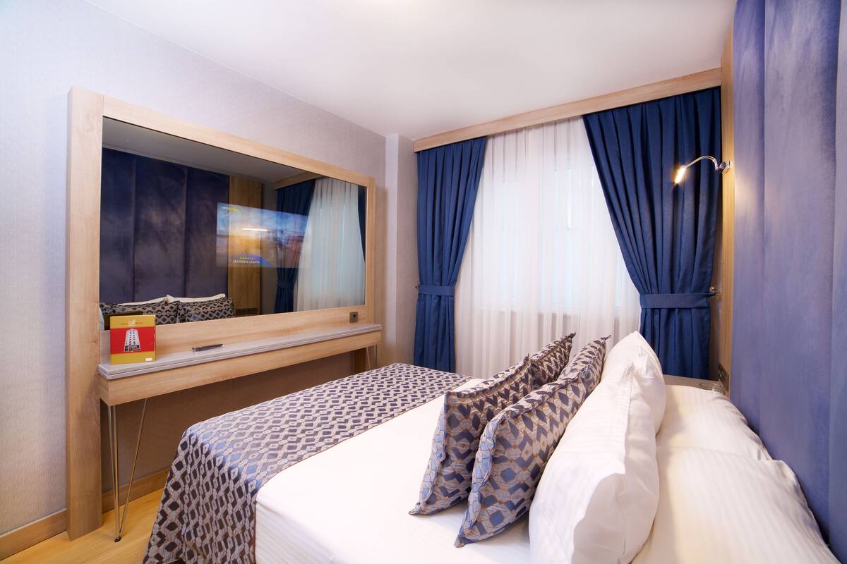 A hotel room with a large bed, a wooden desk, a mirror, and a window with blue curtains.