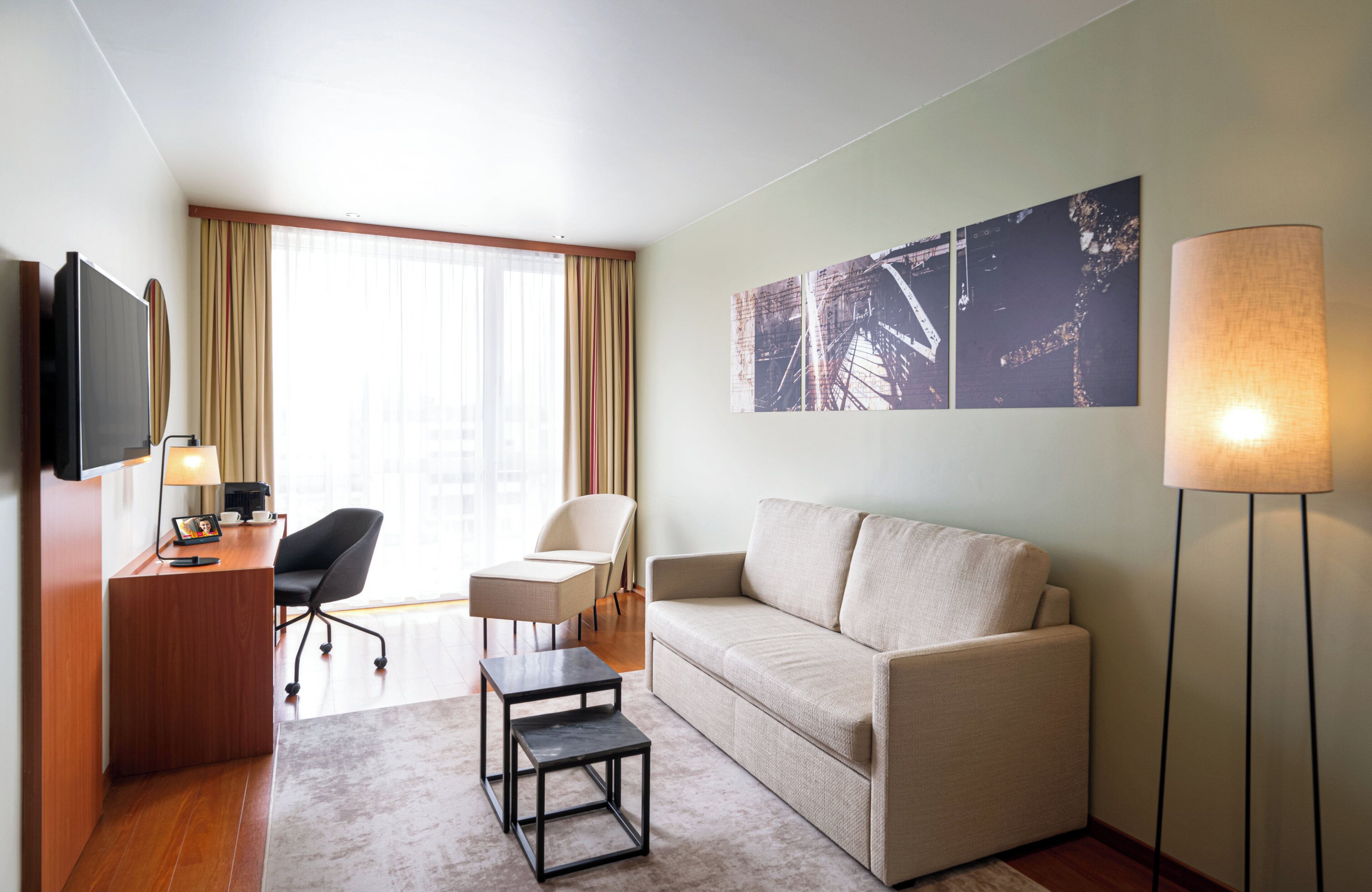 A hotel room with a wooden desk, a sofa, a chair, a small table, a TV, and artwork on the wall.