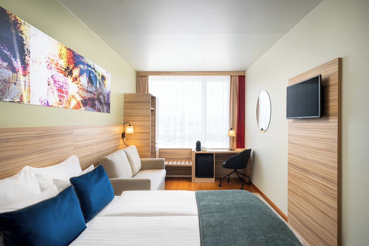 Une chambre d’hôtel avec un lit, un bureau, une chaise, une télévision et un grand tableau abstrait accroché au mur.