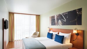 A hotel room with a large bed, two bedside tables, a chair, a TV, and a cityscape painting on the wall. - Leonardo Hotel Salzburg Airport (Wals-Siezenheim)