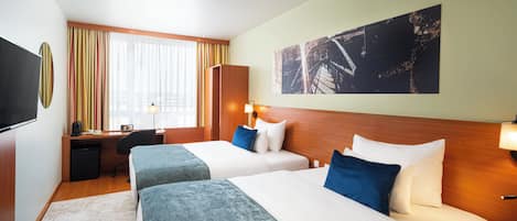 A hotel room with two beds, a desk, a TV, and a large window with curtains.