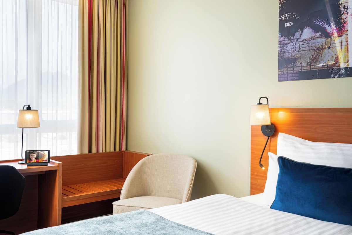 Une chambre d’hôtel avec un lit, un bureau avec une lampe, une chaise et un téléviseur fixé au mur.