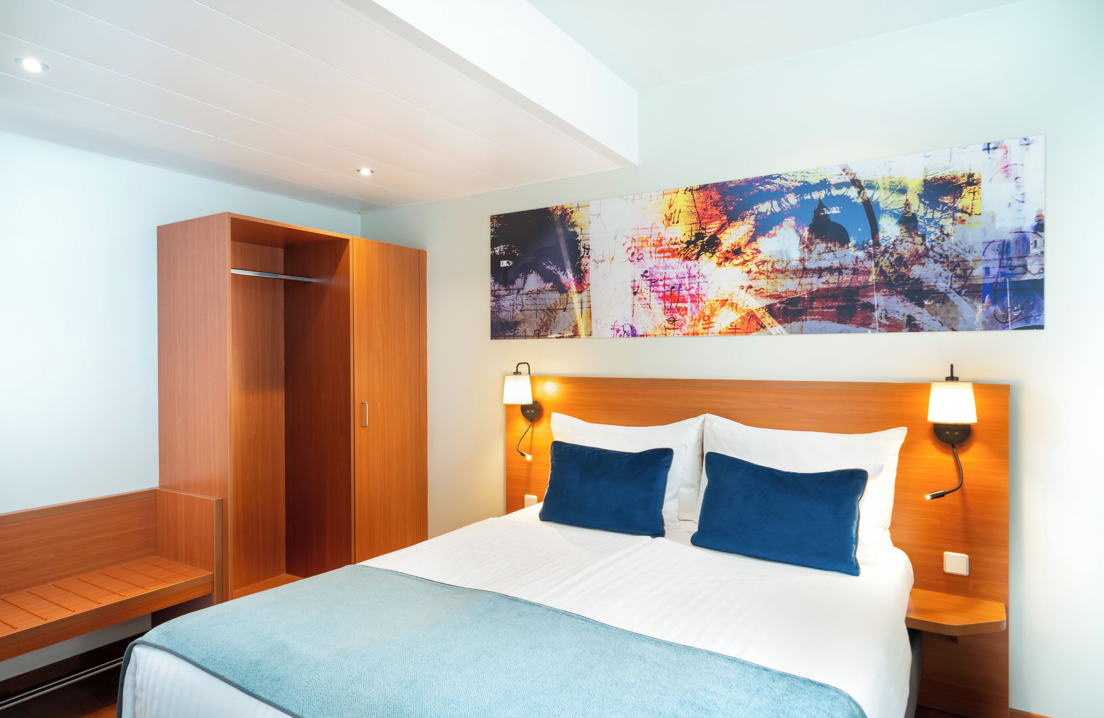 Ein Hotelzimmer mit einem großen Bett, einem Holzschrank, einer Bank und einem an der Wand hängenden, abstrakten Gemälde.