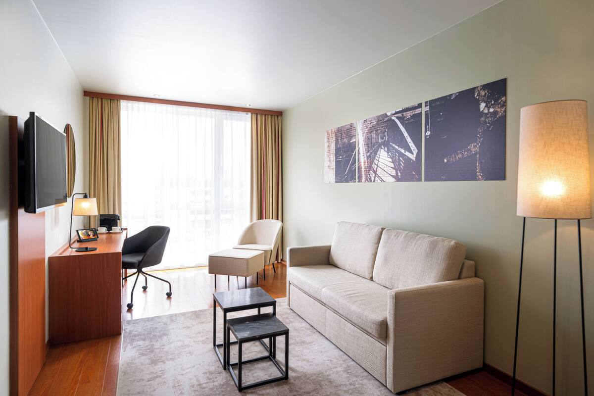 Une chambre d’hôtel avec un bureau en bois, un canapé, une chaise, une petite table, un téléviseur et des œuvres d’art accrochées au mur.