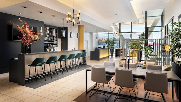 A modern bar area with a long counter, multiple bar stools, and a large decorative plant. - Leonardo Hotel Salzburg Airport (Wals-Siezenheim)