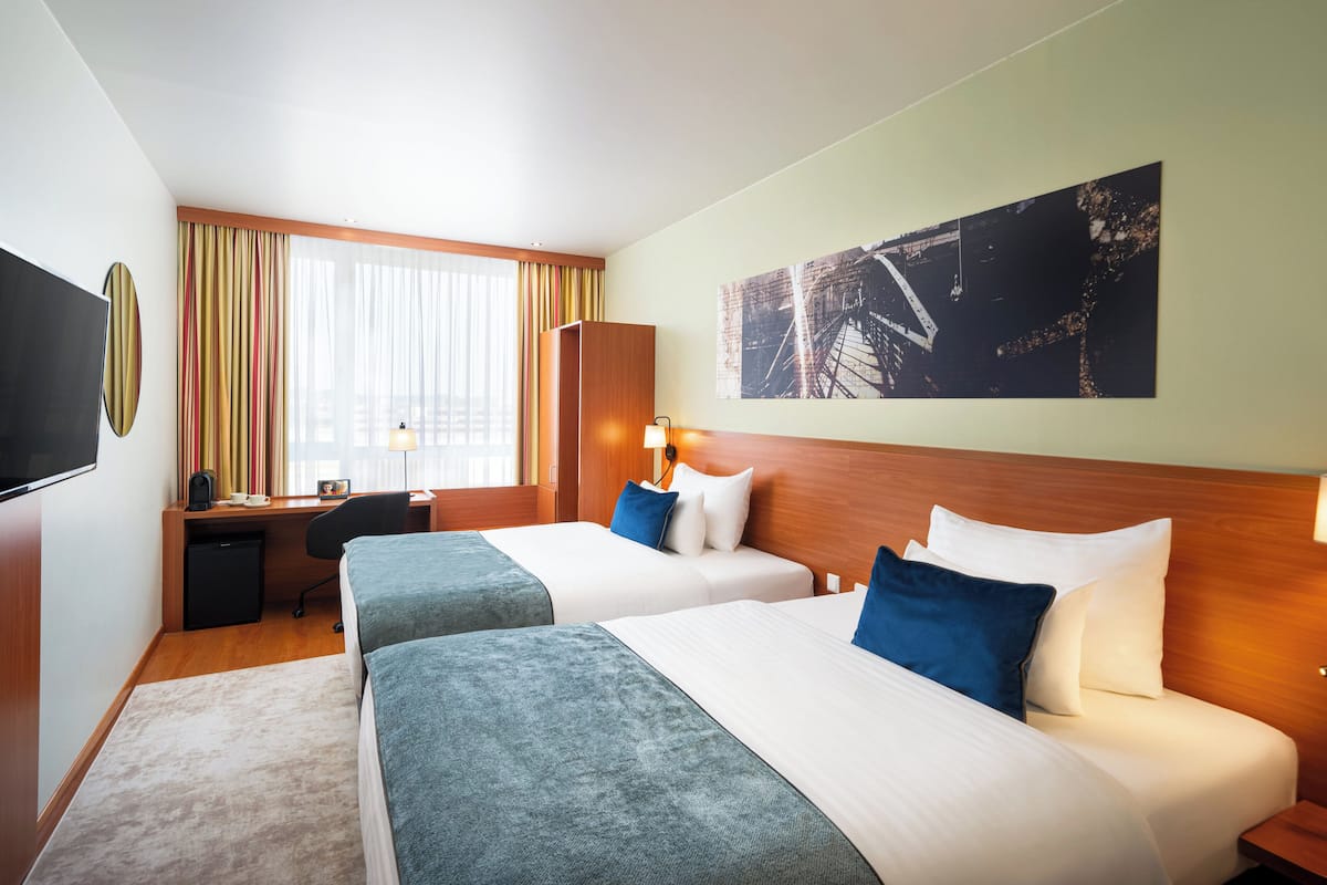 Une chambre d’hôtel avec deux lits, un bureau, une télévision et une grande fenêtre avec des rideaux.