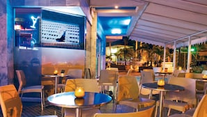 An outdoor seating area with round tables and chairs, a bar with a neon sign, and a view of palm trees and a building in the background. - Sol Caribe Sea Flower (San Andrés)