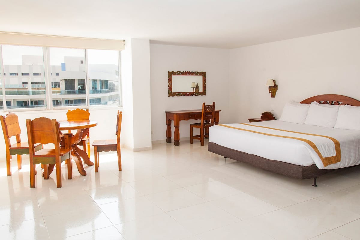 A hotel room with a large bed, a dining table with chairs, a desk, a mirror, and a window with a view of buildings.