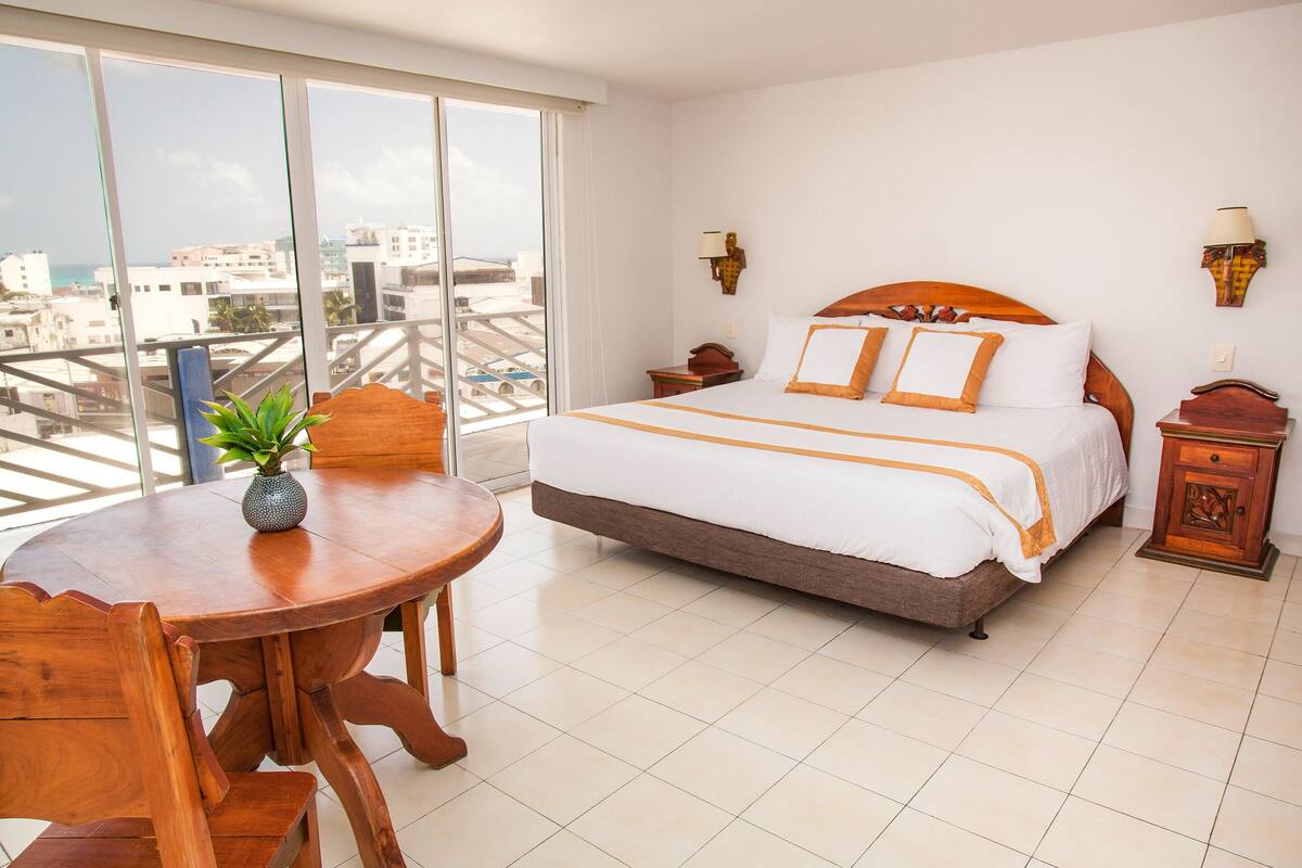 A hotel room with a large bed, a wooden table with a plant, and a view of the city through large windows.