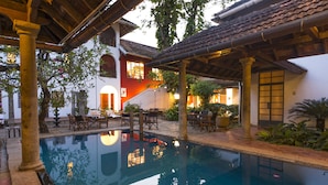 Zona de piscina con suelo de baldosas, sillas de madera y una estructura de pérgola.