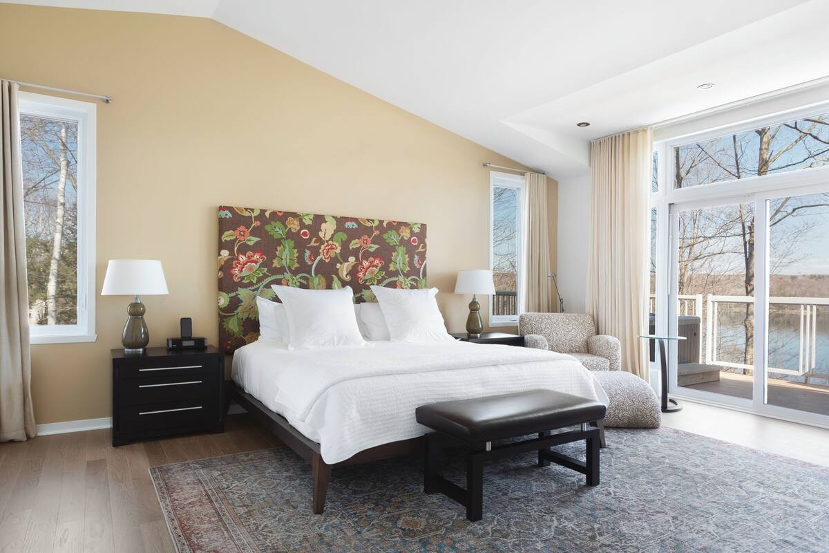 Quarto com uma cama grande, mesa de cabeceira, cadeira, banqueta e uma janela ampla com vista.