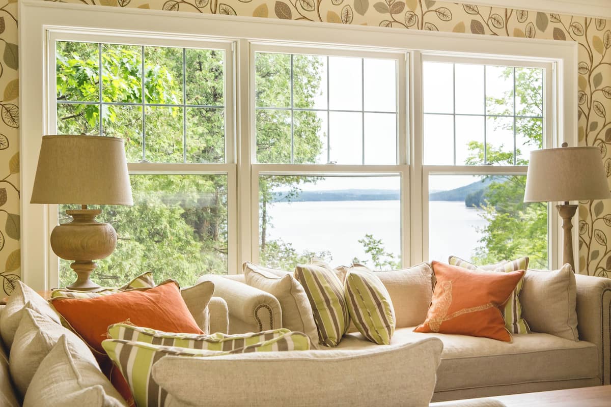 Quarto com grande janela que oferece vista para um lago e árvores. Há um sofá com várias almofadas e duas luminárias de chão.