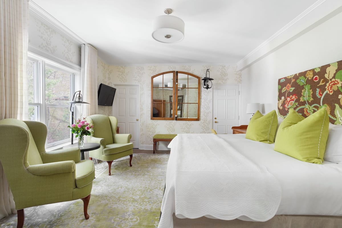 Quarto com uma cama grande, duas poltronas, uma mesinha de centro com um vaso de flores, um espelho e uma televisão na parede.