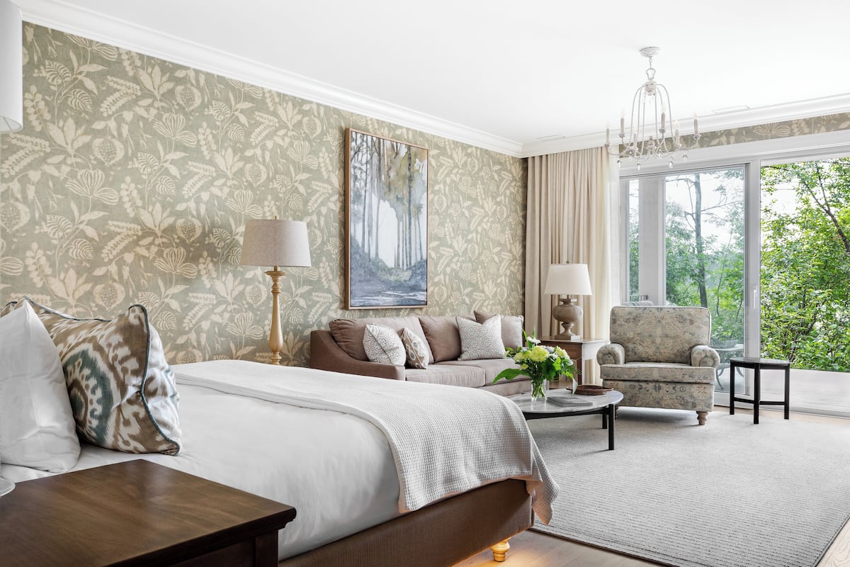 Quarto de hotel com uma cama grande, um sofá, uma poltrona, uma mesa de centro e vista para o verde lá fora.