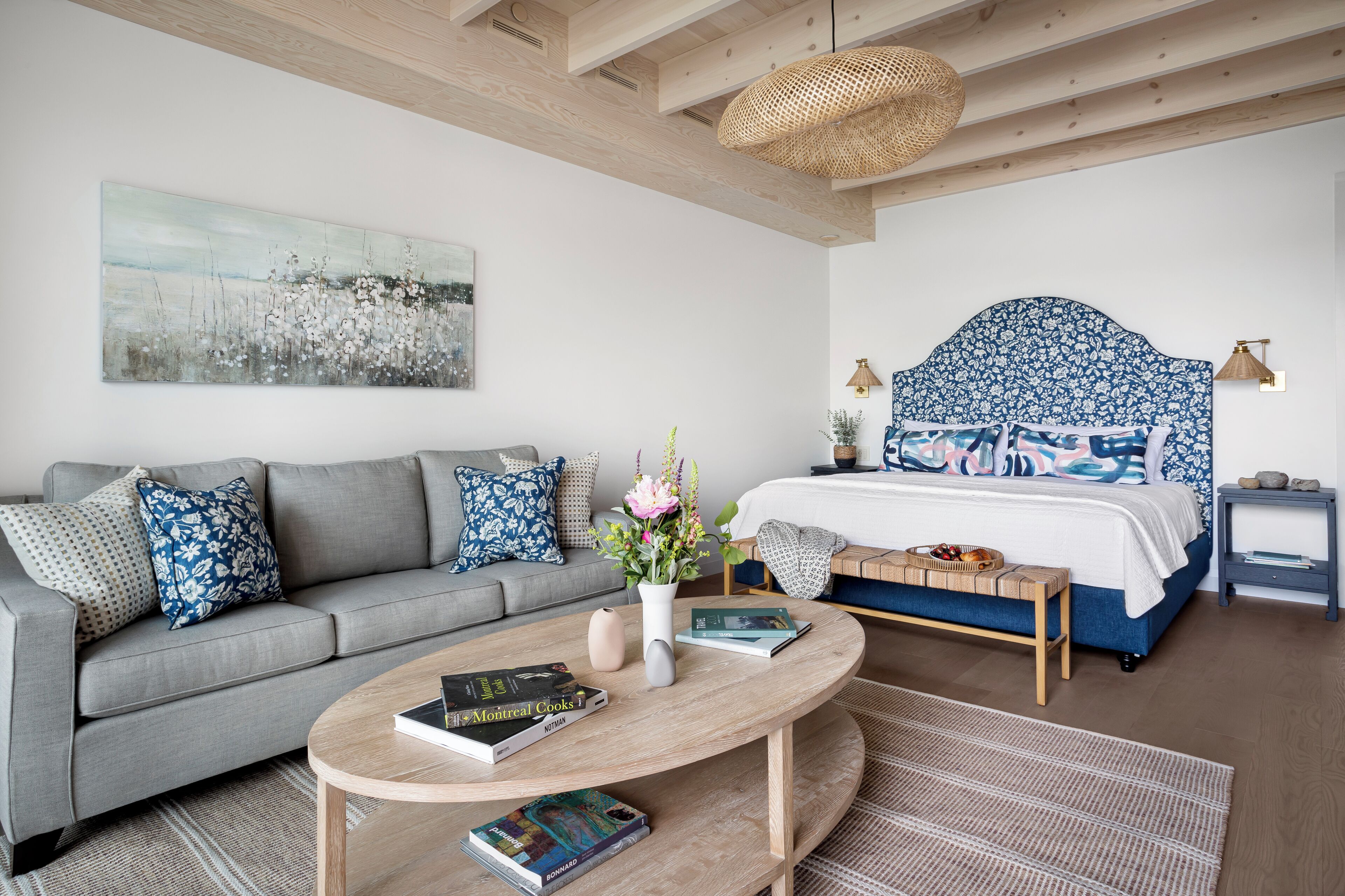A modern bedroom with a grey sofa, a round wooden coffee table, a bed with a blue floral headboard, and a large abstract painting on the wall.