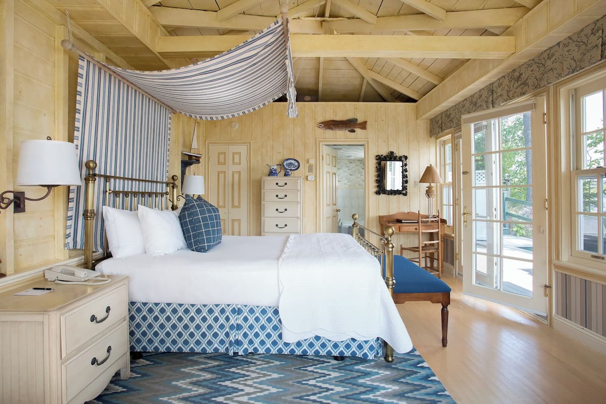Quarto com cama, abajures, escrivaninha e cadeira. Janela com vista para árvores.