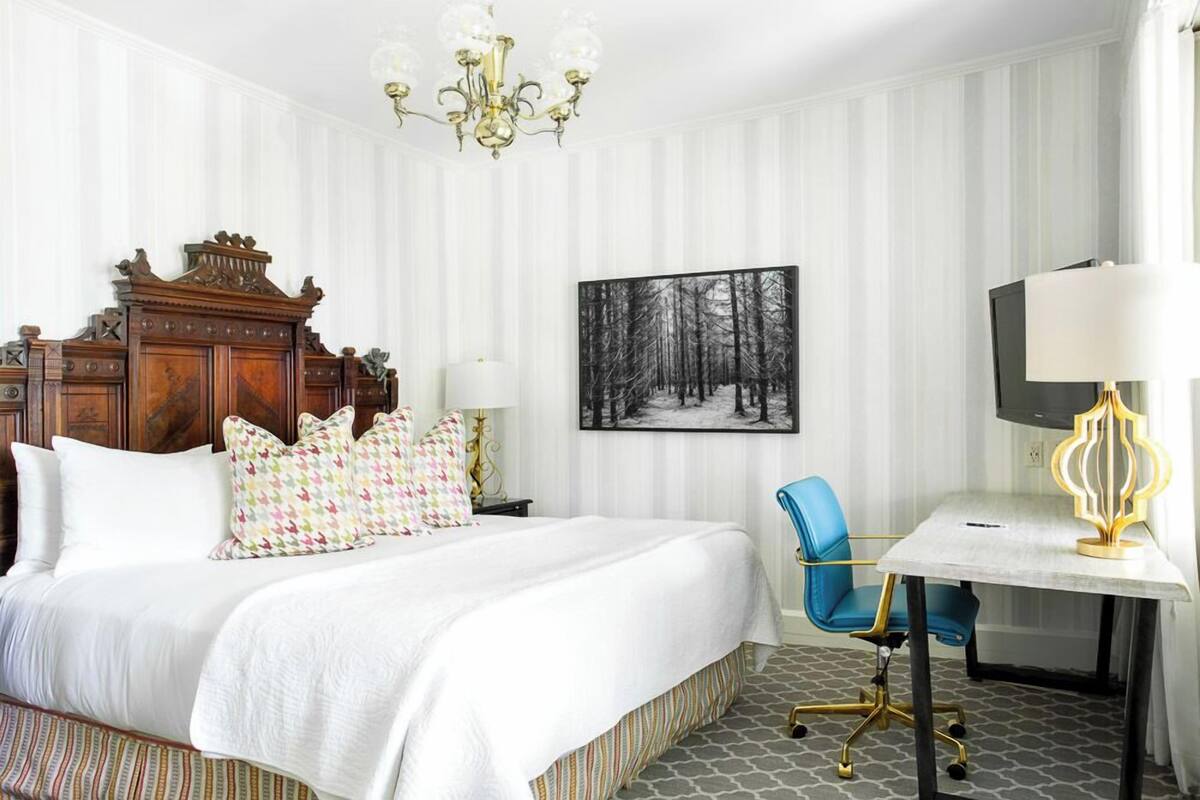 Quarto de hotel com uma cama grande, uma escrivaninha com cadeira, um candelabro e um quadro na parede.
