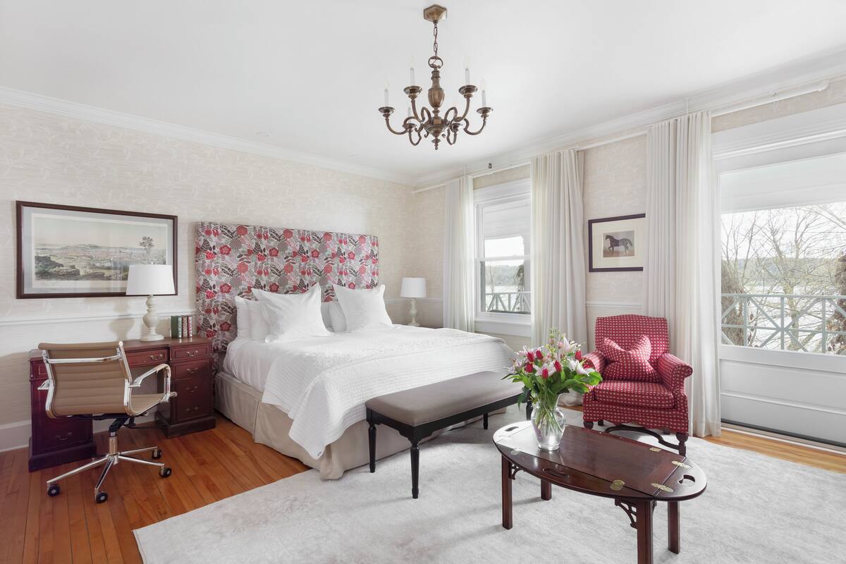 Quarto de hotel com uma cama grande, uma escrivaninha com cadeira, uma poltrona vermelha e um lustre.