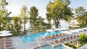 Un área de piscina con sillones de descanso y sombrillas, rodeada de árboles y con vista a un cuerpo de agua.