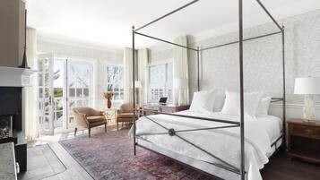 A spacious bedroom with a four-poster bed, a fireplace, a desk with a laptop, and a view through large windows.
