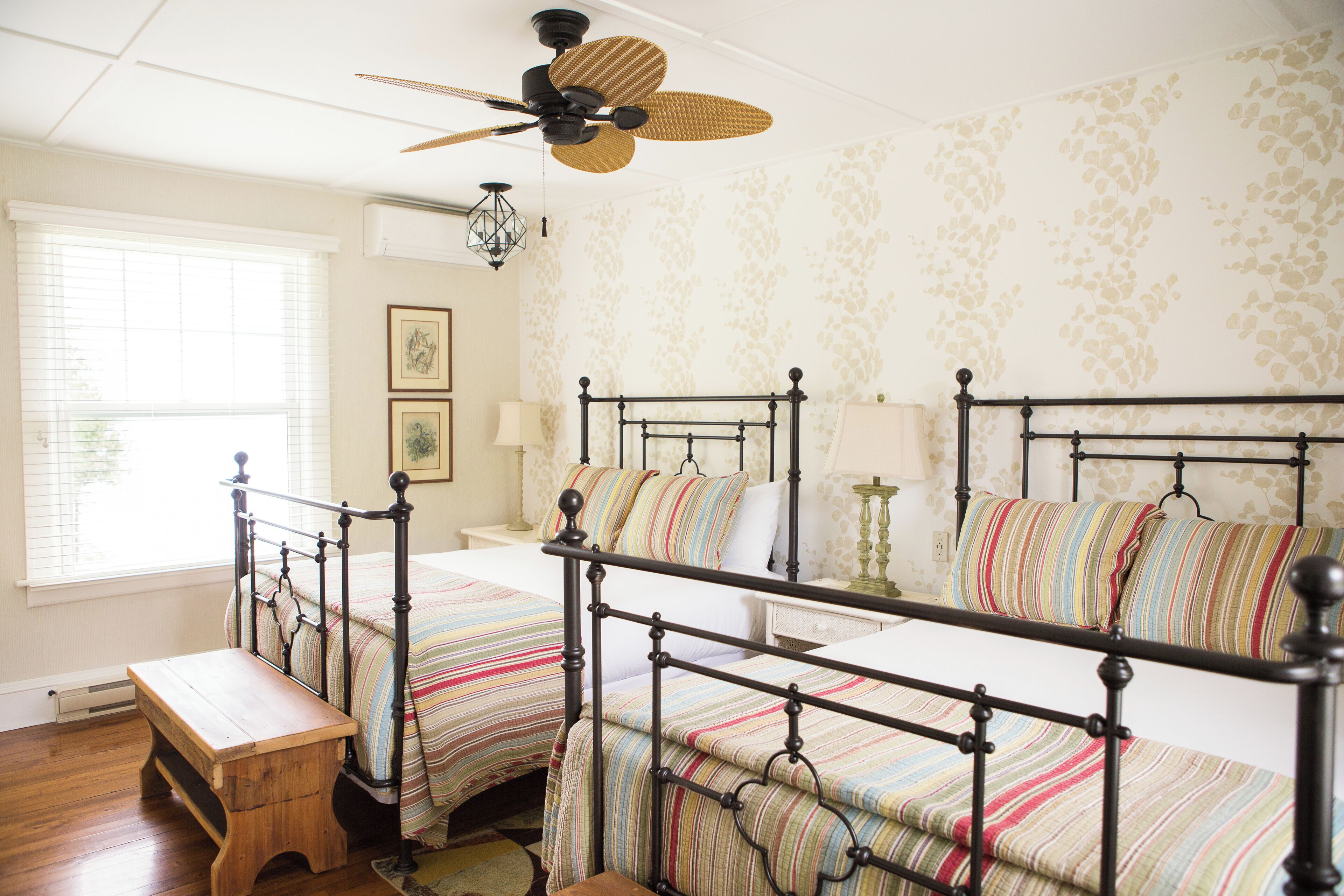A bedroom with two beds, a wooden bench, a ceiling fan, and a window with blinds.