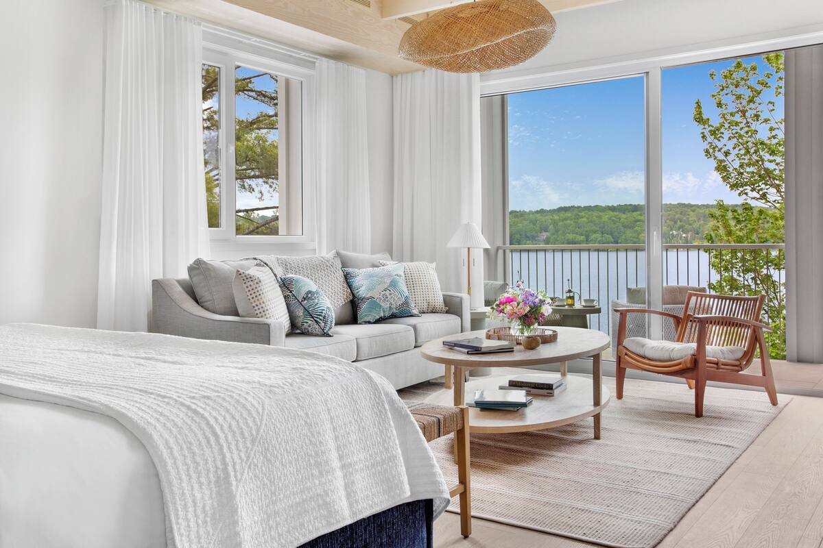 Quarto moderno com uma cama grande, um sofá, uma cadeira, uma mesa de centro redonda e vista para um lago através de amplas janelas.