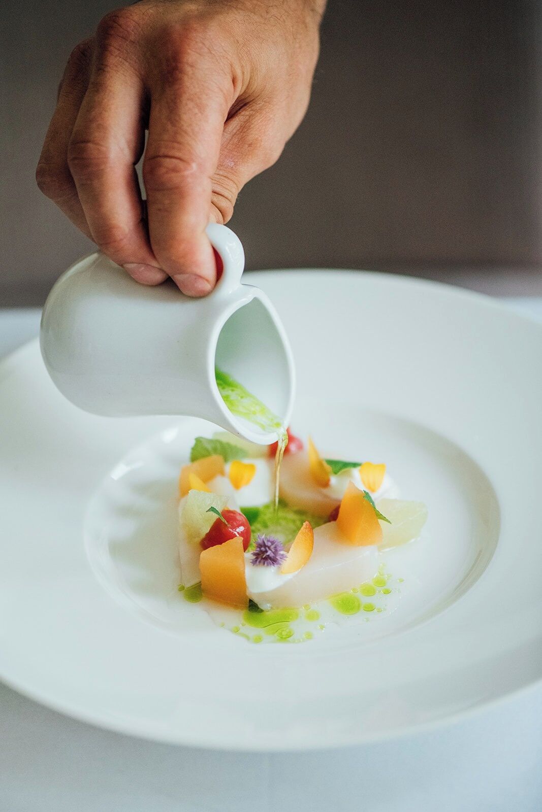 A hand pouring a green sauce onto a plate with a white sauce and fruit garnish.