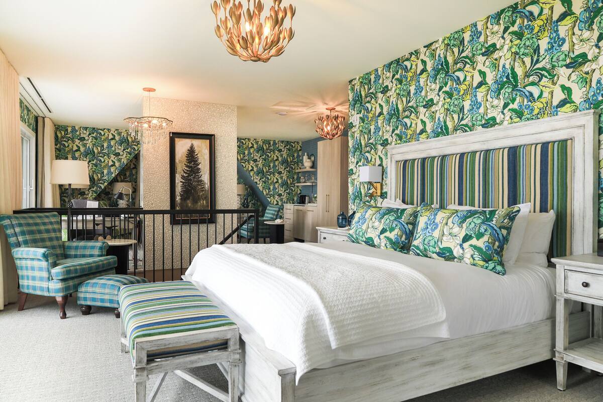 Quarto com uma cama grande, uma cadeira, um banco, uma mesa de cabeceira e um candelabro.