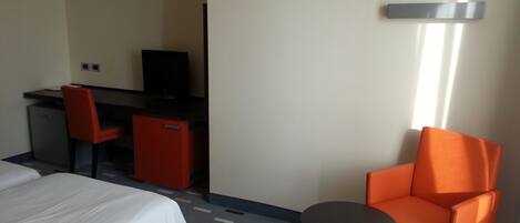 Una habitación de hotel con una cama, un escritorio con una computadora, una mesita pequeña y una silla naranja.
