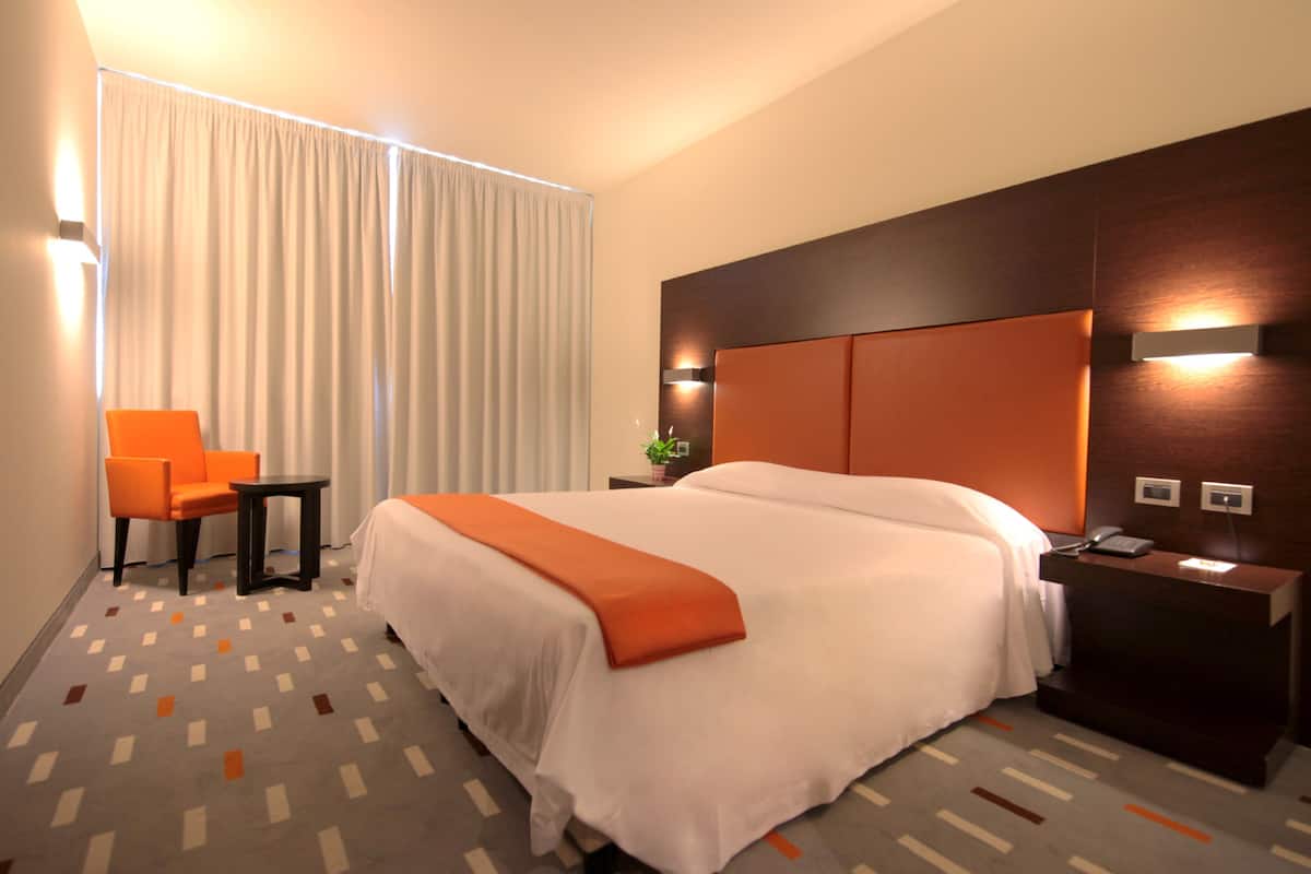 Habitación de hotel con una cama grande, un sillón naranja, una mesita pequeña y una mesita de noche con un teléfono.