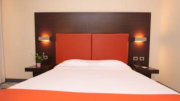 A hotel room with a large bed, two bedside tables, a nightstand, a potted plant, and wall-mounted lighting.