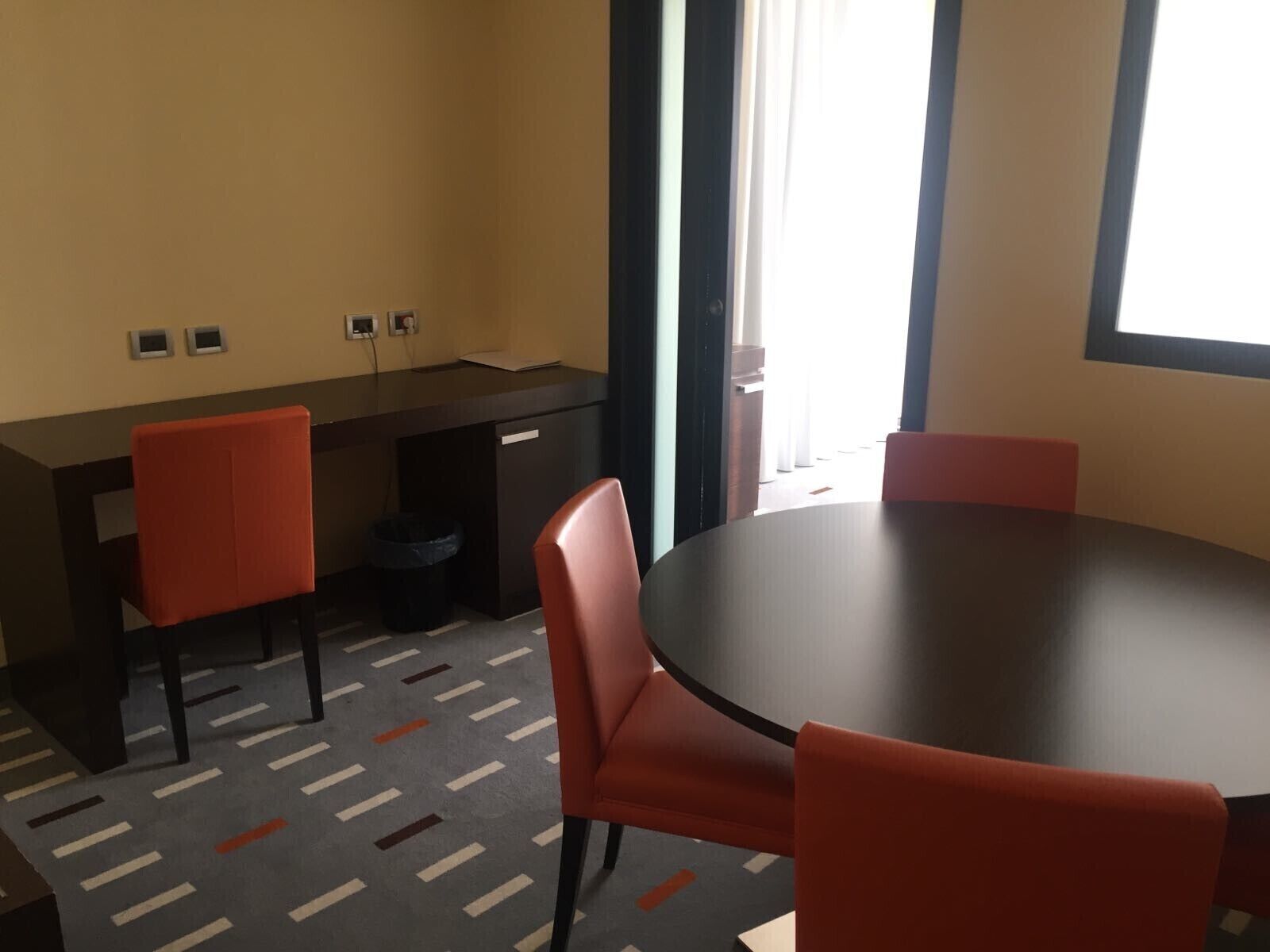 A conference room with a round table, four chairs, a desk, and a trash bin.