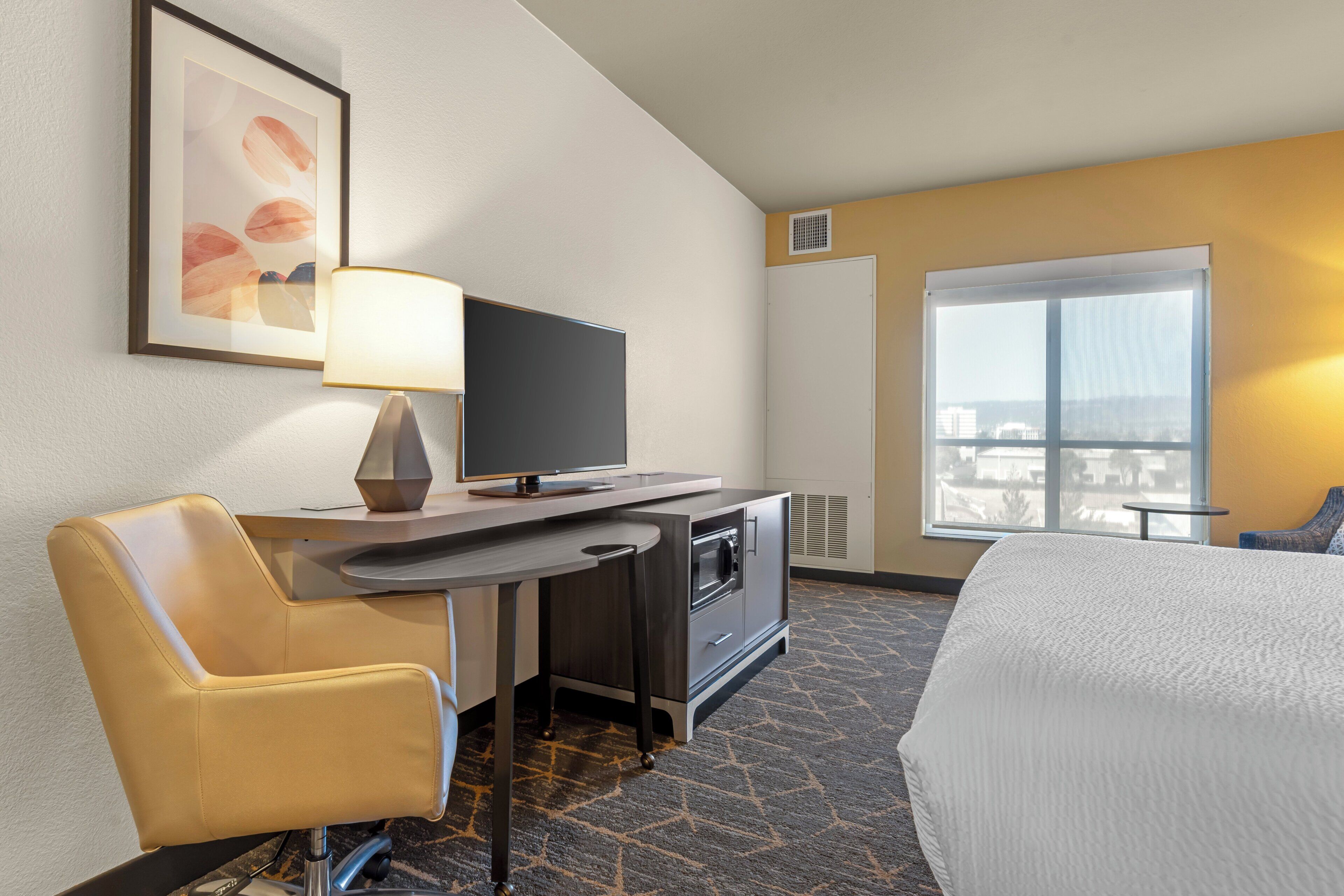 Ein Hotelzimmer mit Bett, Schreibtisch mit Computer, Stuhl, Fernseher und Fenster mit Blick auf die Stadt.