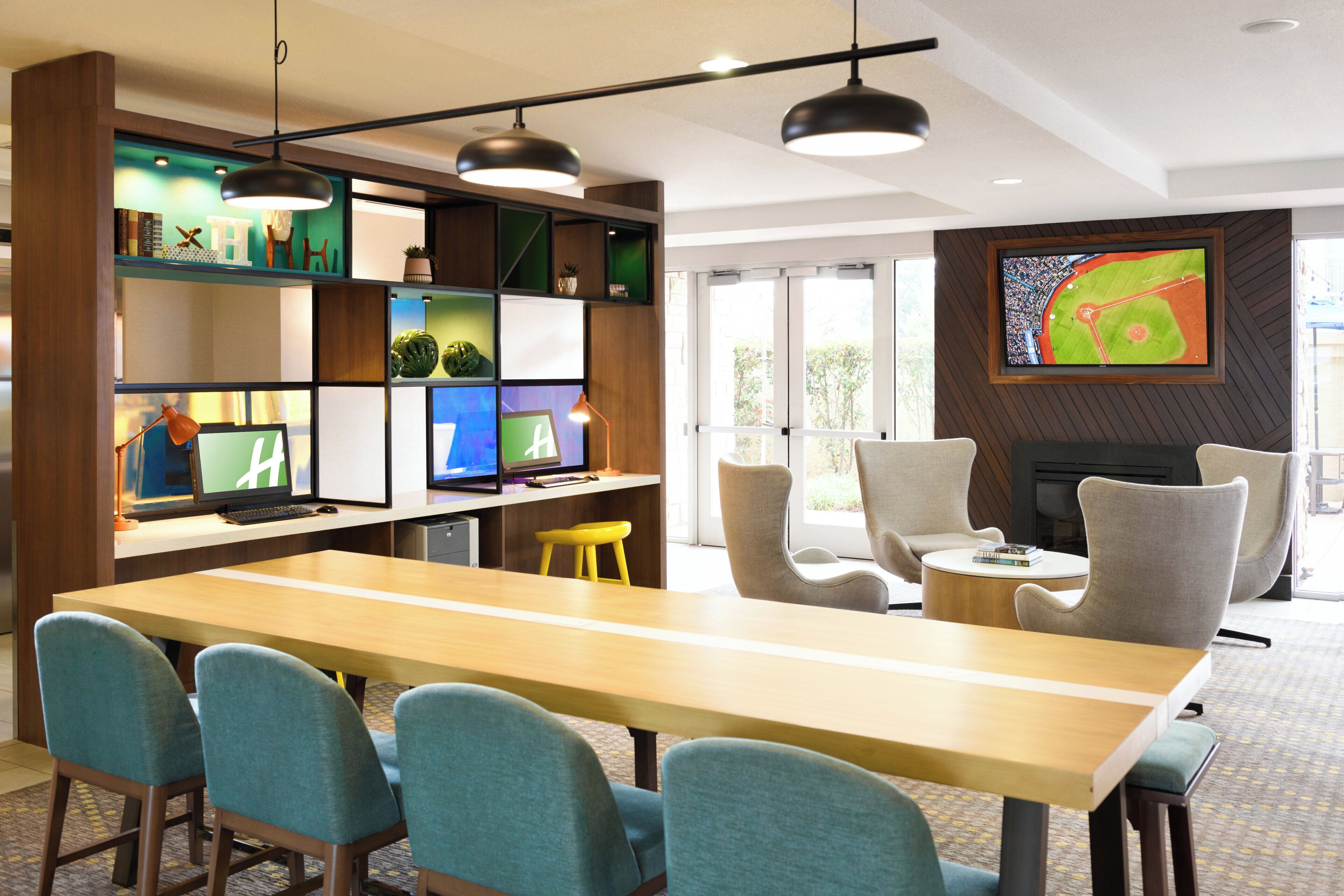 Habitación de hotel moderna con una larga mesa de madera, varias sillas, un televisor montado en la pared y una estantería con diversos objetos.