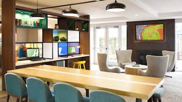 Habitación de hotel moderna con una larga mesa de madera, varias sillas, un televisor montado en la pared y una estantería con diversos objetos.