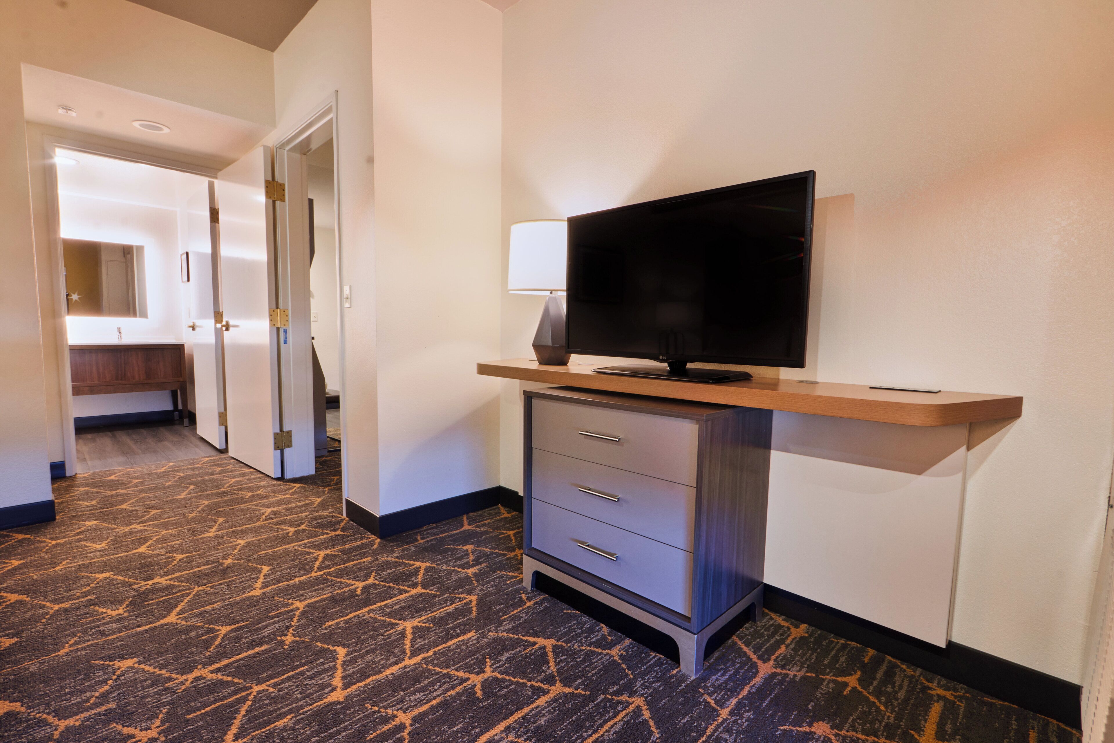 Un pasillo de hotel con suelo alfombrado, un televisor sobre un soporte, un escritorio con cajones y un espejo en la pared.