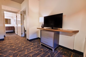 Un pasillo de hotel con suelo alfombrado, un televisor sobre un soporte, un escritorio con cajones y un espejo en la pared.