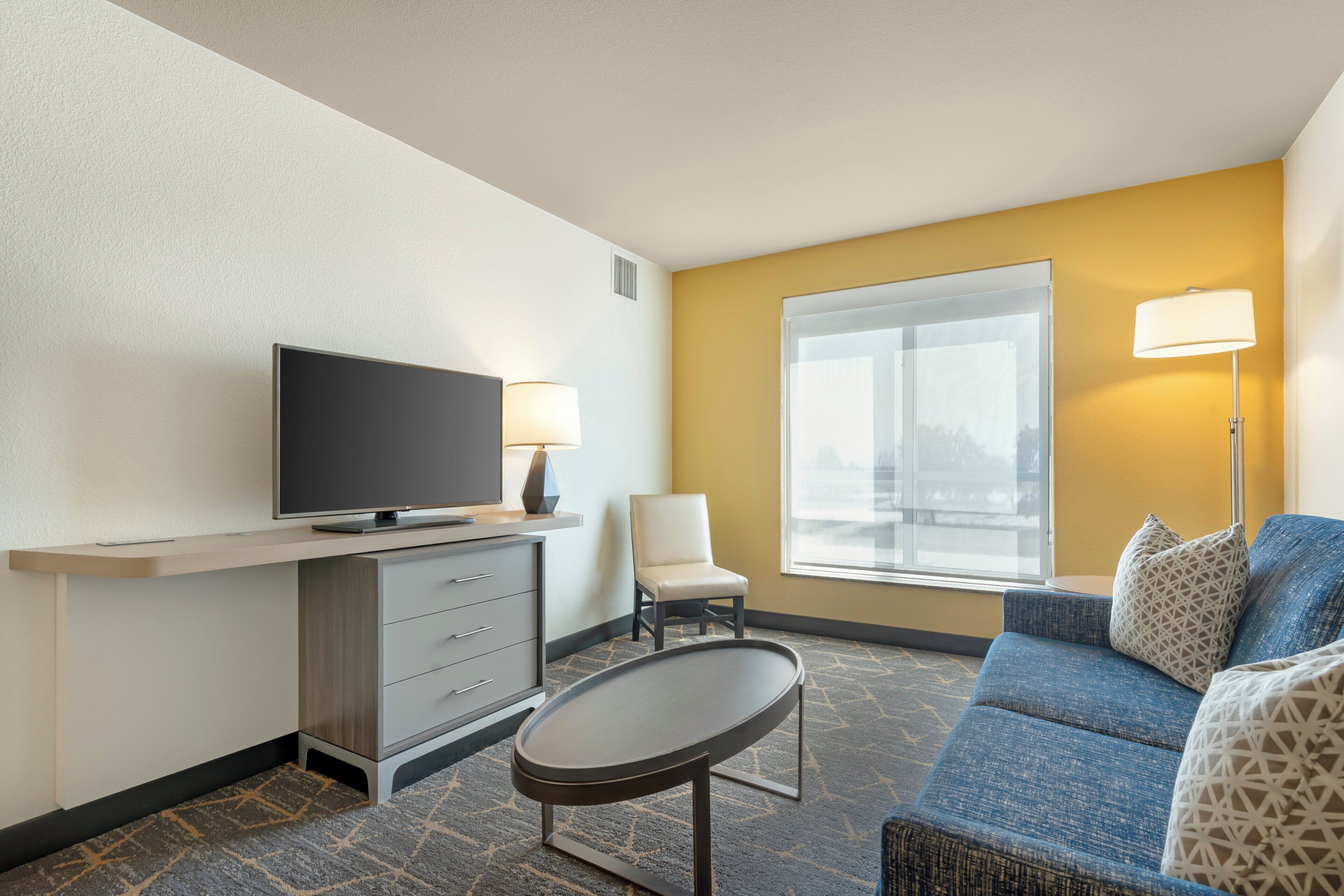A hotel room with a television on a desk, a chair, a sofa, and a window with a view.