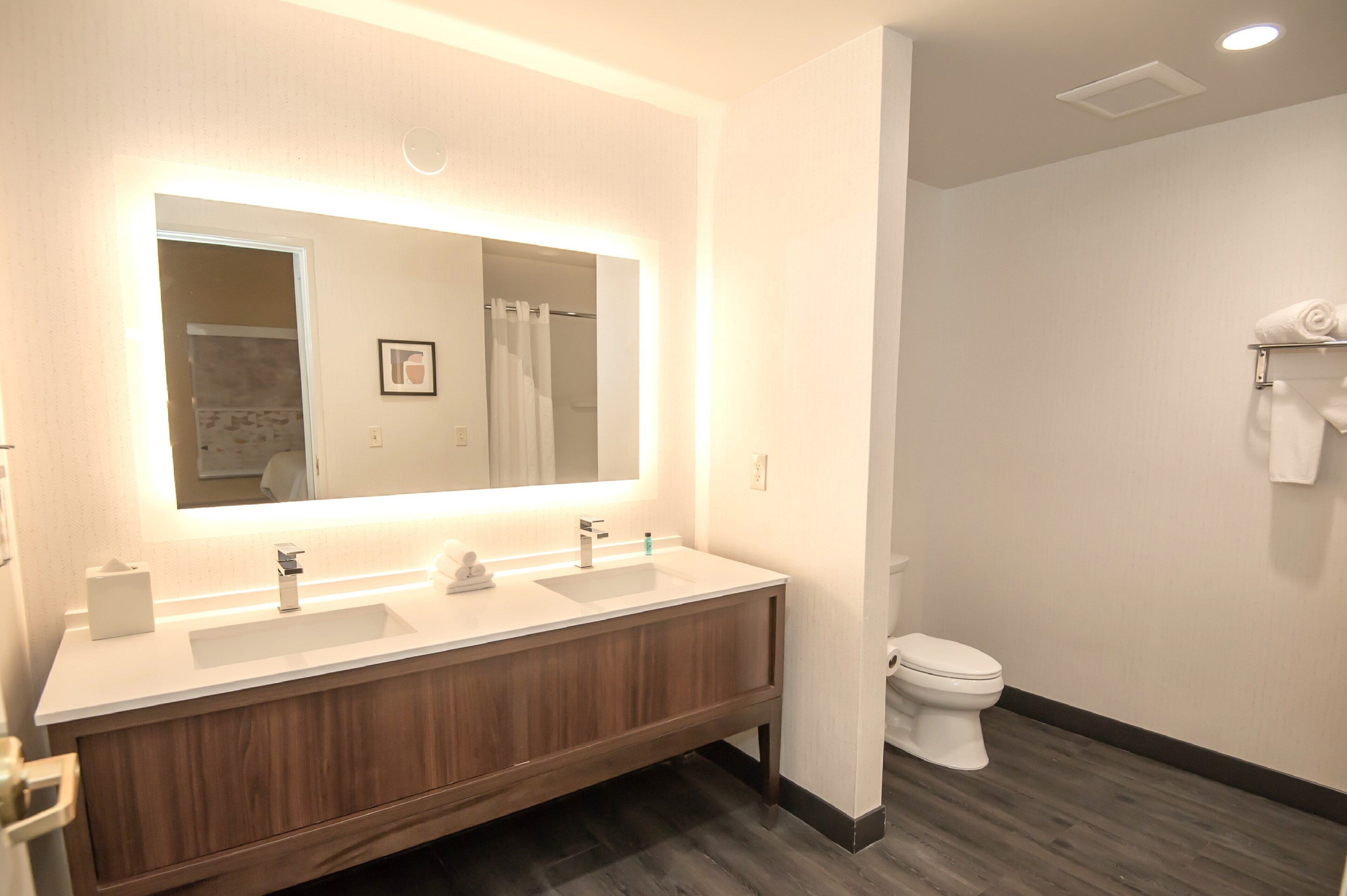 A modern bathroom with a large mirror, two sinks, a toilet, and a shower area.