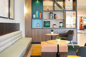 Un área de descanso moderna con un sofá, dos mesas, una estantería y un detalle decorativo en la pared.