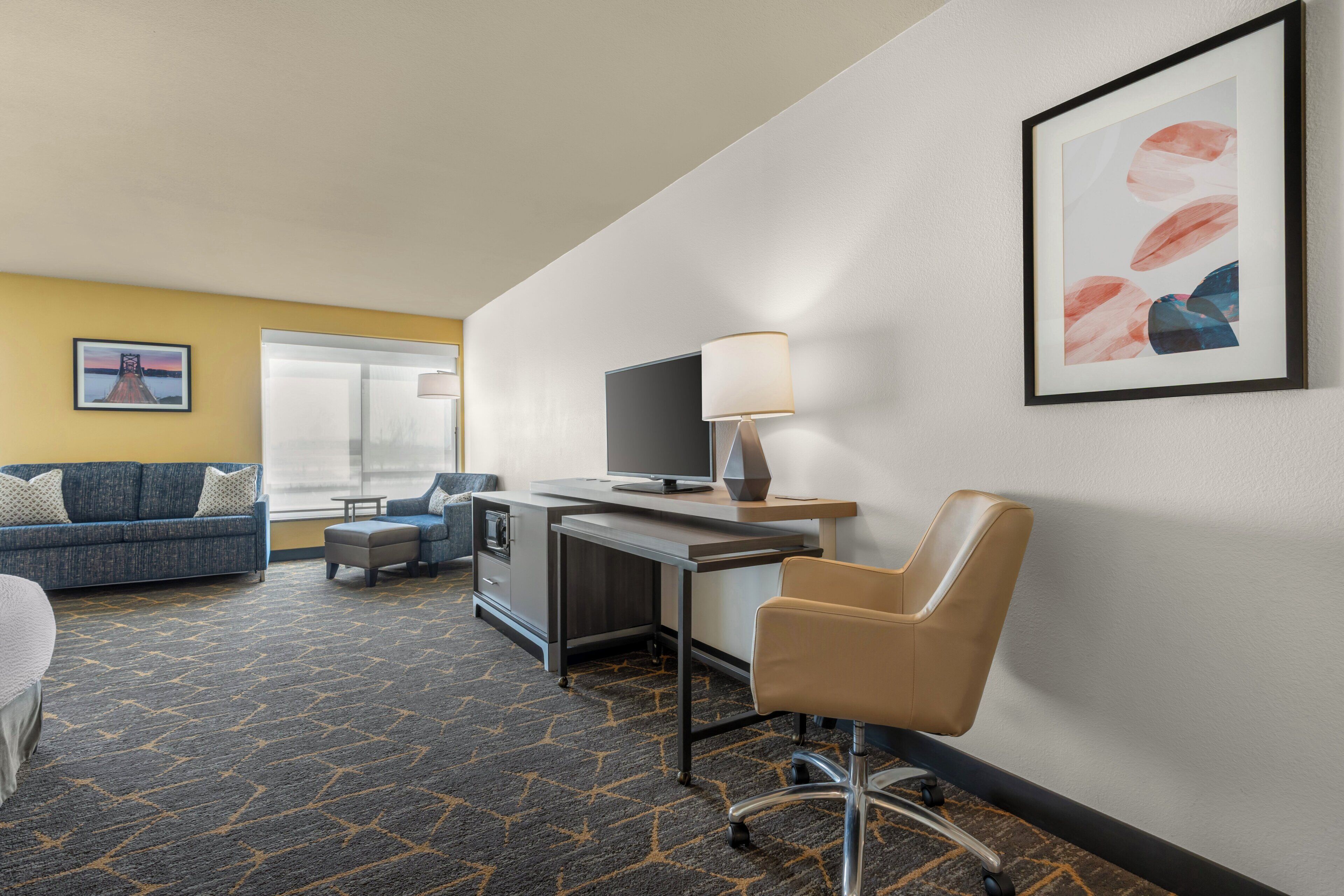 A hotel room with a sofa, a desk, a chair, a television, and a window.