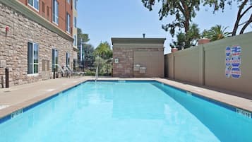 A swimming pool with a stone wall and a building on one side, and a fenced area with trees and a sign on the other.