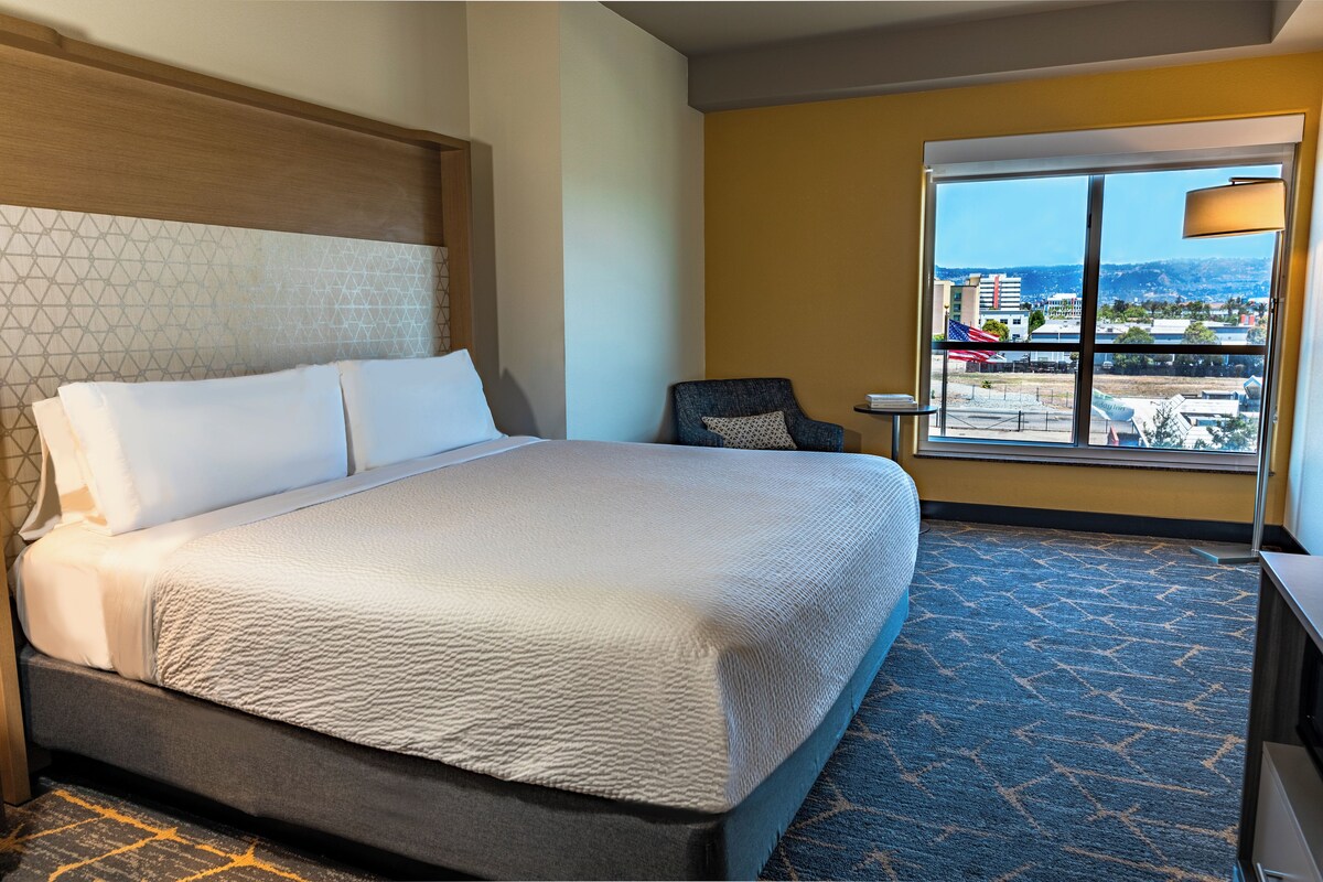 Quarto de hotel com uma cama grande, uma cadeira, uma mesinha e uma janela com vista para a cidade.