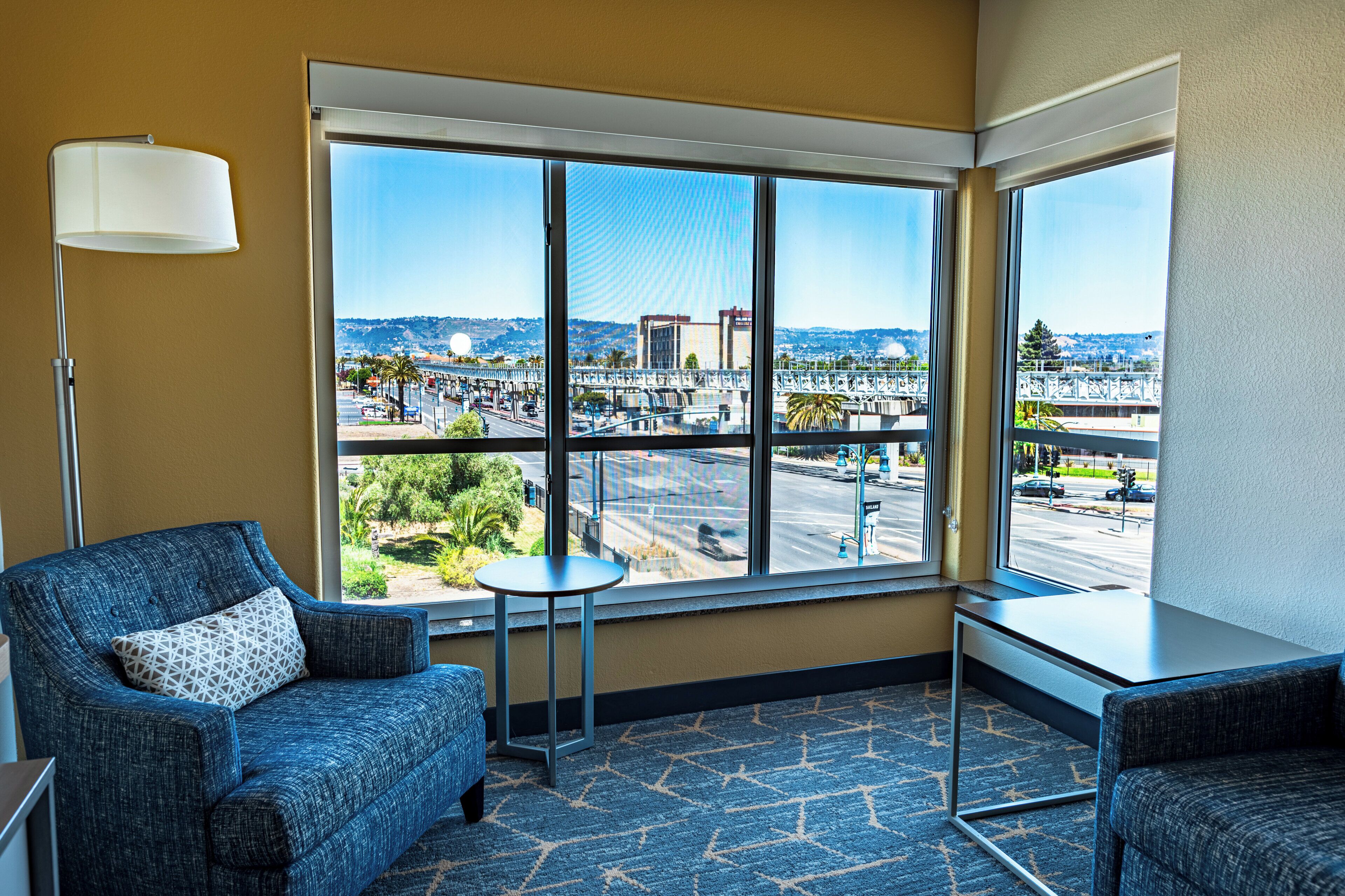 A hotel room with a large window offering a view of a cityscape, a blue armchair, a small table, and a lamp.
