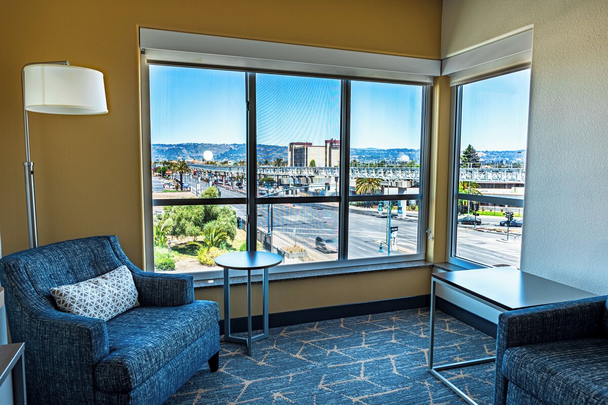 A hotel room with a large window offering a view of a cityscape, a blue armchair, a small table, and a lamp.