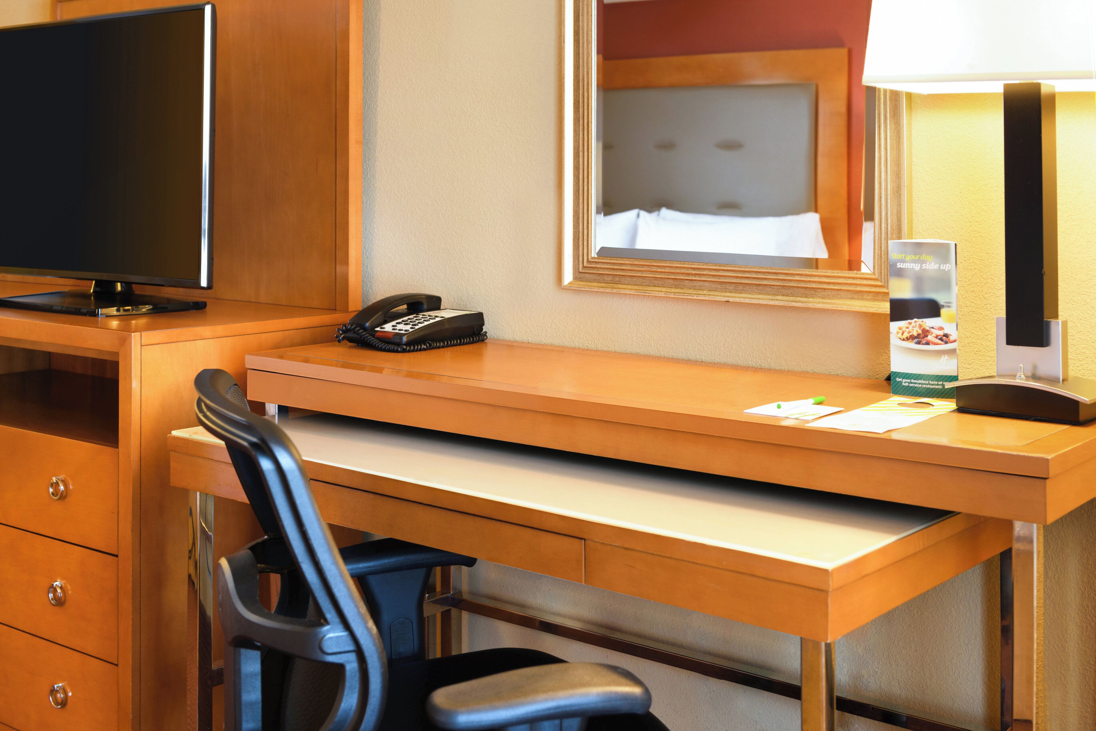 A hotel room desk with a flat-screen TV, a telephone, a mirror, and a lamp.