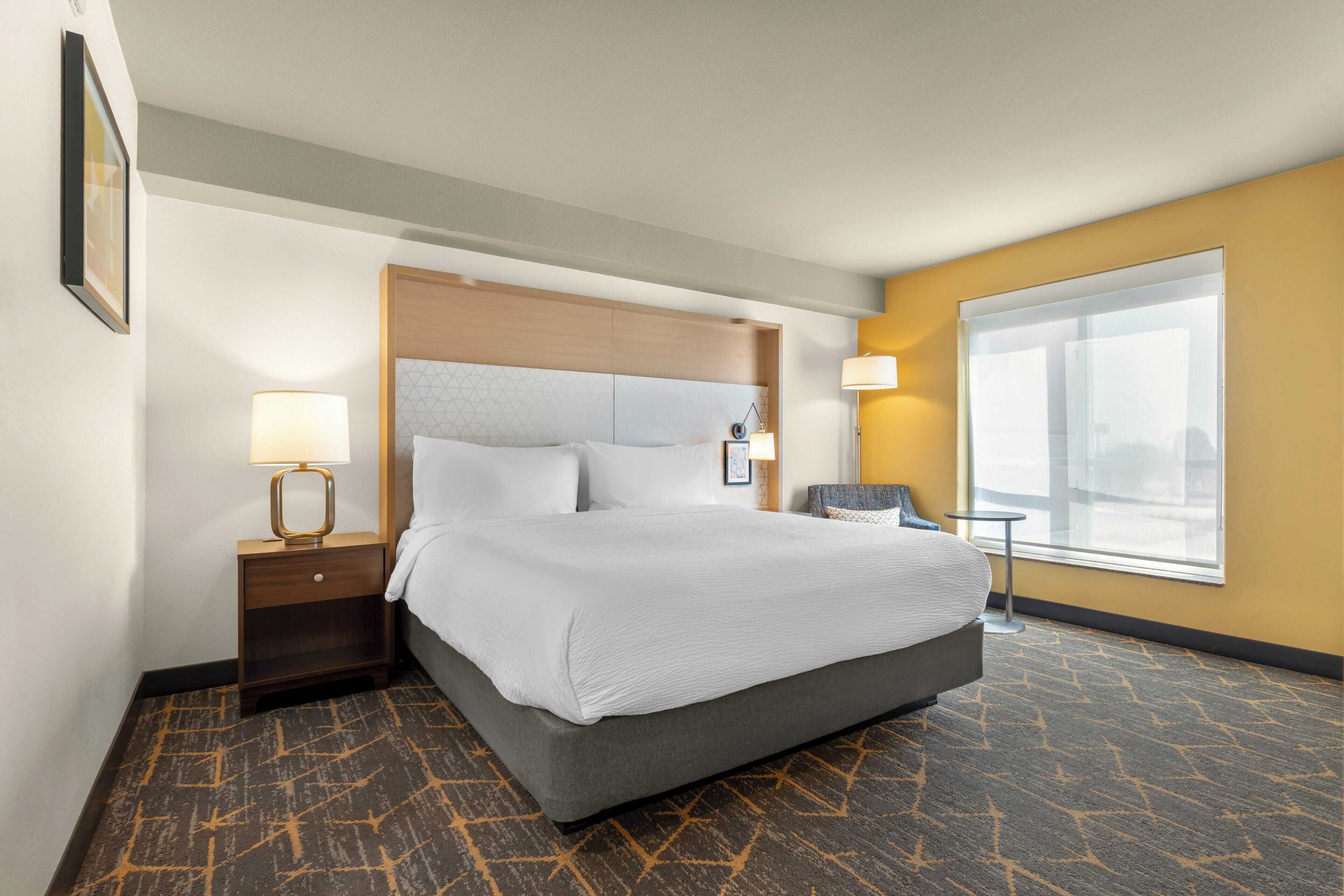 Une chambre d’hôtel avec un grand lit, une table de chevet, une chaise, une fenêtre et une lampe.