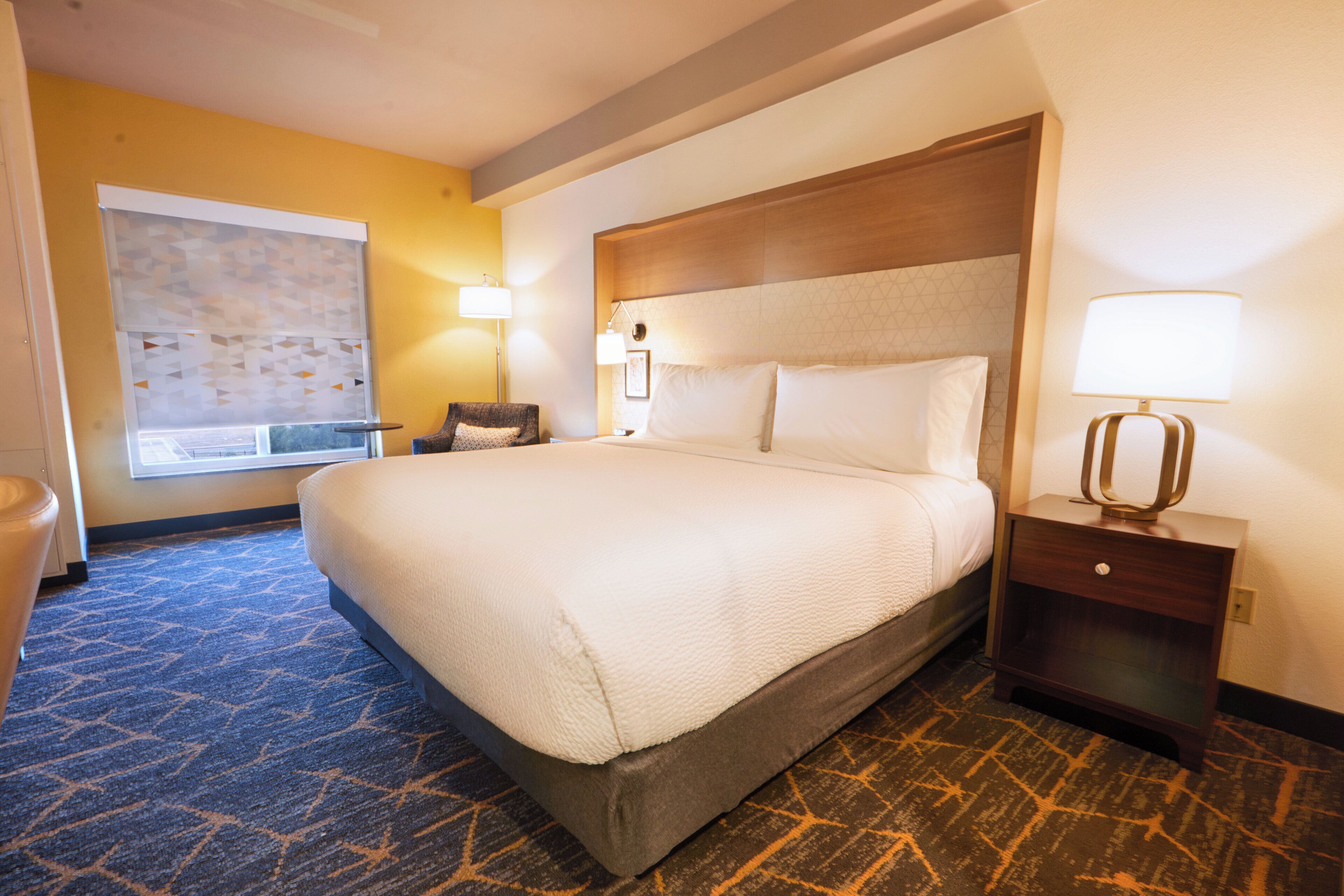 A hotel room with a large bed, a nightstand with a lamp, a chair, and a window with blinds.